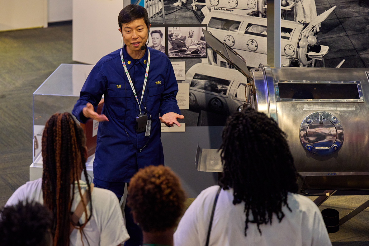 person speaking to a group in front of an iron lung