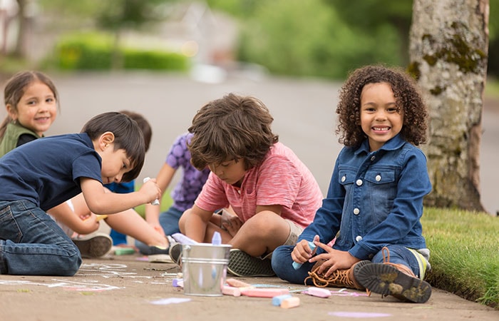 A photo of a young child sitting on the ground smiling with chalk in their hand as friends in the background play with chalk on pavement.