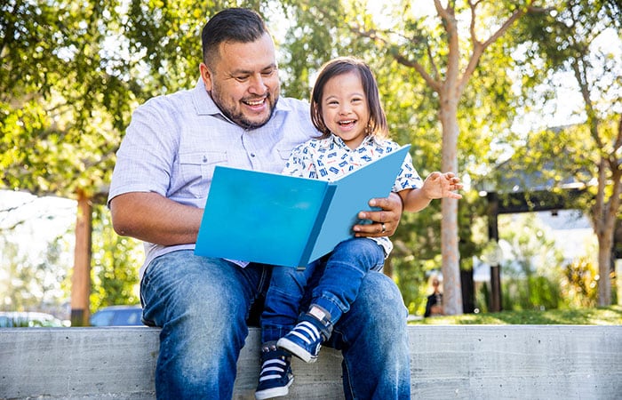 Padre hispano leyéndole un libro al aire libre a su hijo que tiene síndrome de Down sentado en las piernas