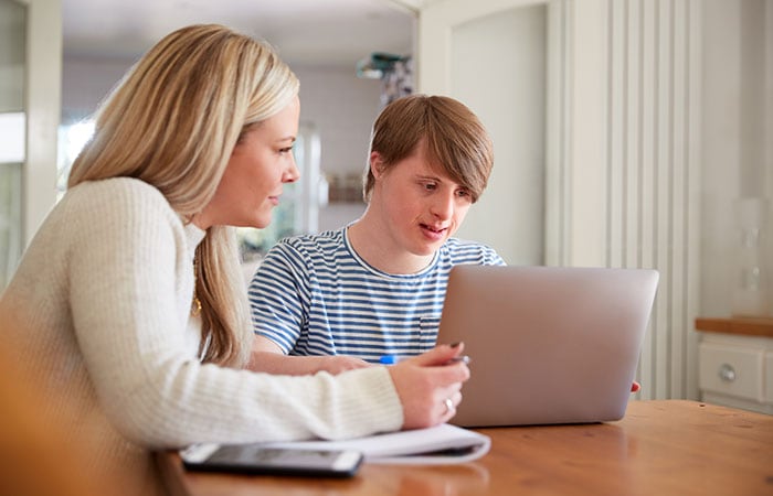 Madre caucásica sentada a la mesa de la cocina con su hijo adolescente con síndrome de Down que usa su computadora portátil.