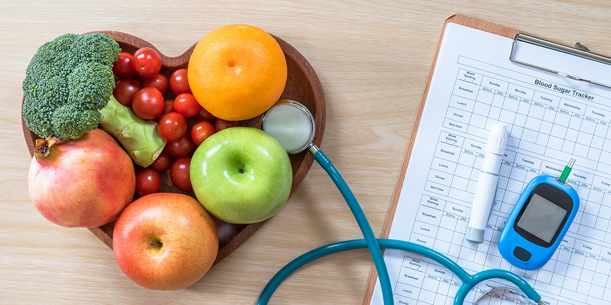 A heart shaped-bowl with colorful fruits and vegetables sits on a table with a stethoscope, glucose monitor, and clipboard.