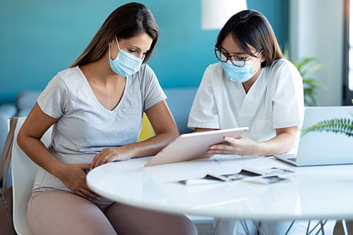 Young beautiful gynecologist wearing a hygienic face mask while showing to pregnant woman ultrasound scan baby with digital tablet in medical consultation.