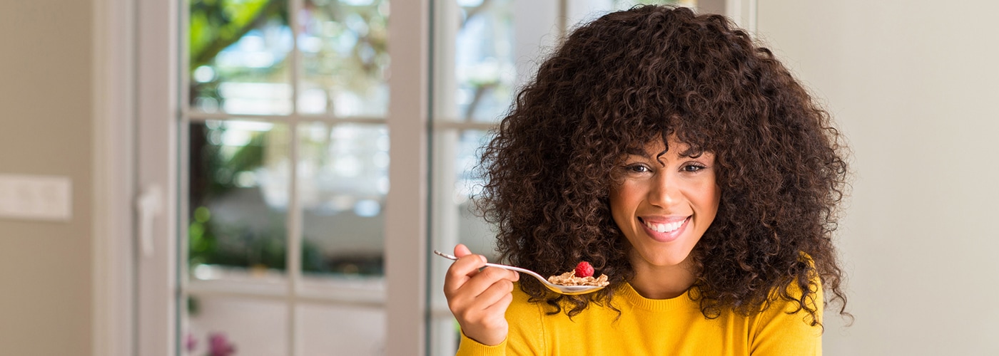 Mujer afroamericana comiendo cereales, frambuesas y arándanos con una cara feliz de pie y sonriendo con una sonrisa segura mostrando dientes