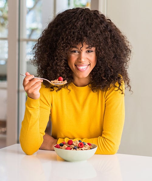 African american woman eating cereals, raspberries and blueberries with a happy face standing and smiling with a confident smile showing teeth