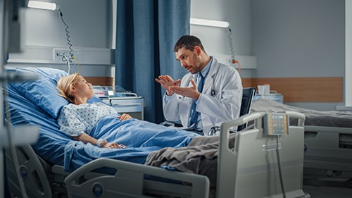 An emergency medicine physician in a white lab coat talking to a patient in a hospital bed.