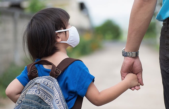 Little girl with backpack wearing cloth face mask hand in her father's hand waiting for school bus. Children need help with the transition back to school.
