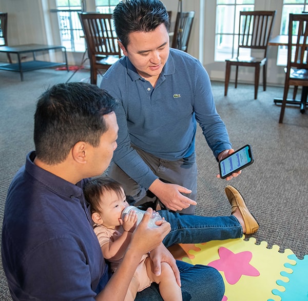 Dads look at the Milestone Tracker app while one dad feeds their baby a bottle.