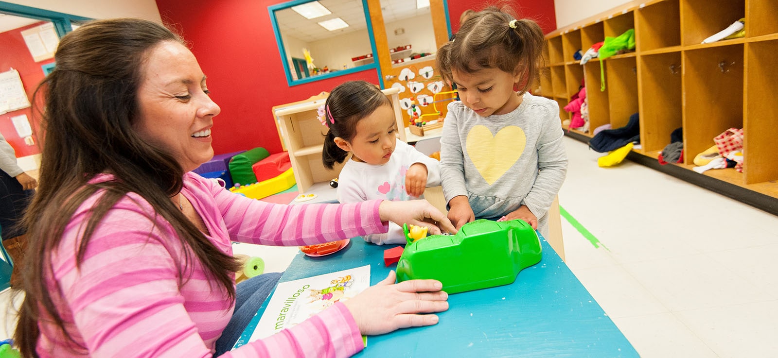 Niñas juegan con juguetes en el salón de clases mientras que una educadora de primera infancia observa, ayuda y sonríe.