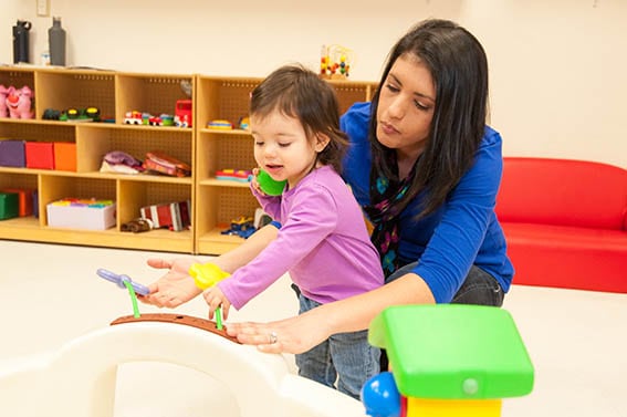 Una niña juega con un juguete en el salón de clases mientras que una educadora de primera infancia la observa por encima de su hombro y la ayuda