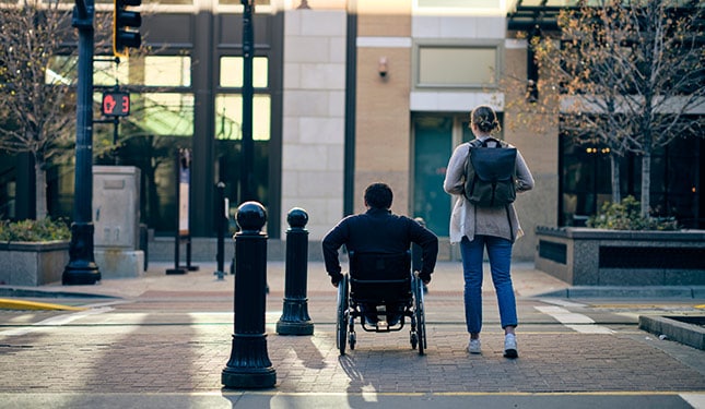 A man in a wheelchair and a friend in a downtown area crosswalk.
