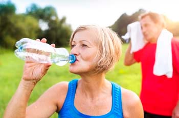 Senior woman drinking from water bottle outdoors with senior man wiping face with towel in background