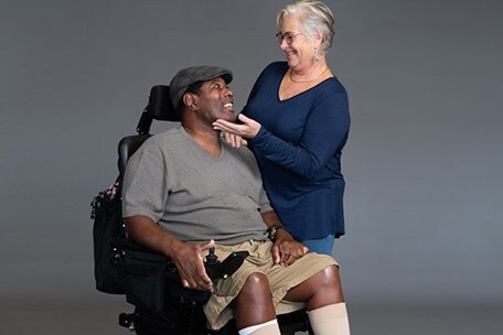 A man with ALS in a wheelchair exchanges a smile with a caregiver.