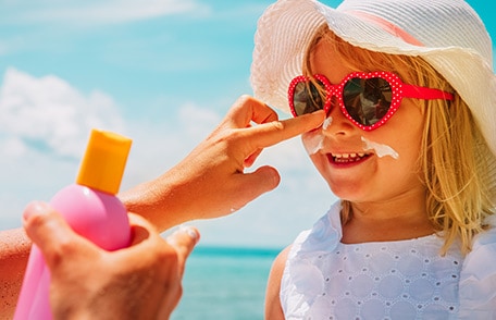 Child wearing a brimmed hat and sunglasses has sunscreen applied to her face.