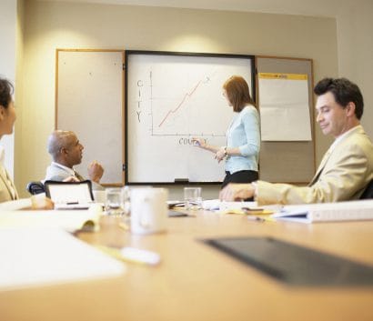 People sitting around a conference table. One is at a white board pointing at chart.