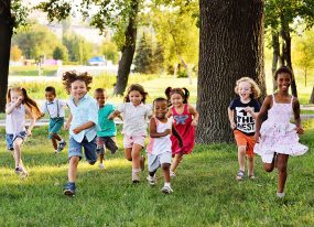 A group of happy children of all ethnicities running together in a grassy park on a sunny day.