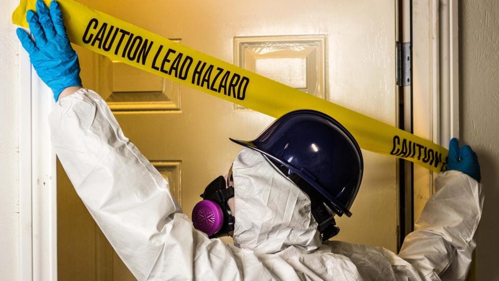 Person in hazmat suit putting tape across a door that reads caution lead hazard.