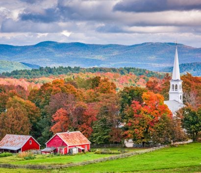 Red barn and white church steeple sticking out of tree-lined hills with fall folliage