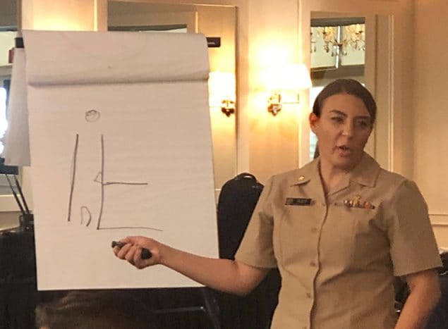 White woman in a beige uniform pointing to a white board and speaking to an audience.