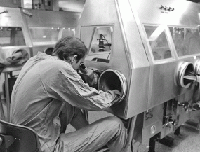 Dr. Russell Regnery manipulates a microscope through airtight glove portals while seated at a Class III cabinet in Building 9 in 1978.