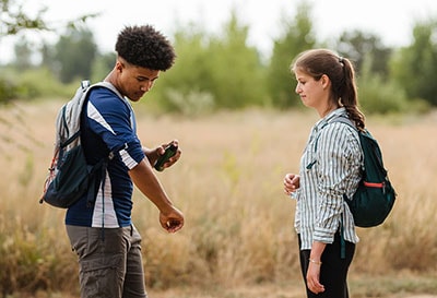 Two young people standing in field while one of them spray insect repellent on his arm.