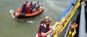 Imagen de una empleada de los CDC subiendo por una escalera para abordar un buque con colaboradores de la Guardia Costera y de la Oficina de Aduanas y Protección Fronteriza de los EE. UU. Al fondo se ve un bote con otras dos personas