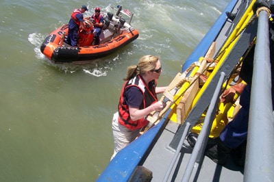 Imagen de una empleada de los CDC subiendo por una escalera para abordar un buque con colaboradores de la Guardia Costera y de la Oficina de Aduanas y Protección Fronteriza de los EE. UU. Al fondo se ve un bote con otras dos personas