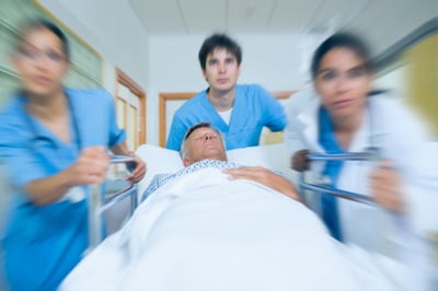 Imagen de tres médicos con batas azules empujando una camilla en la que llevan a un paciente