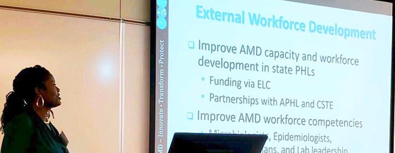 A woman stands to the side of projection screen. The slide reads: External Workforce Development. Improve AMD capacity and workforce. - Funding via ELC - Partnerships with APHL and CSTE. Improve AMD workforce competencies. – Microbiologists, Epidemiologists, Bioinformaticians, and Lab Leadership
