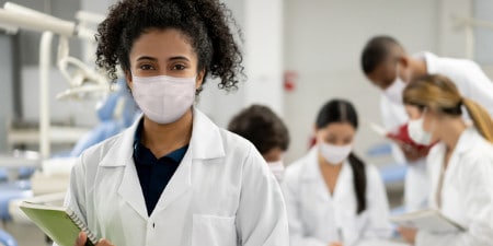 A technician wearing a facial covering in a lab setting with several colleagues meeting in the background