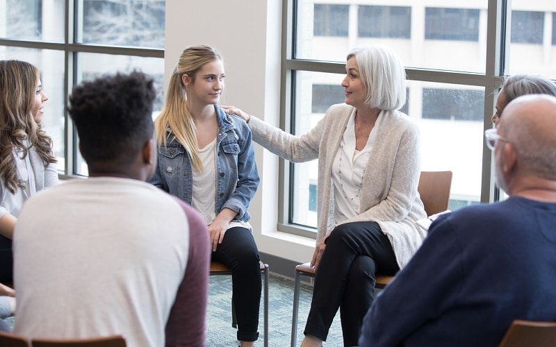 During group therapy, the mature adult woman shares her personal experiences with the teen girl.
