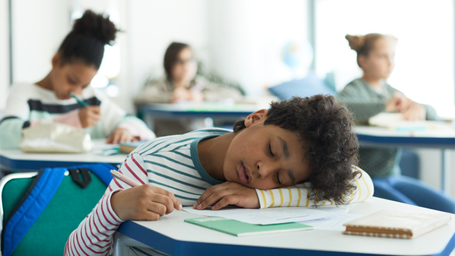 A young boy asleep in a classroom with his head on his desk.