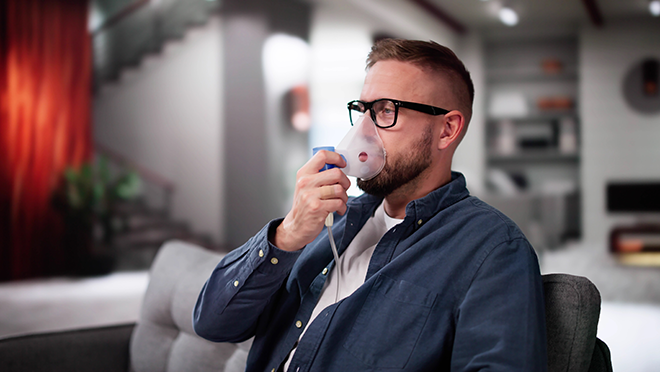 Man sitting on the couch using a breathing mask to treat a breathing condition.
