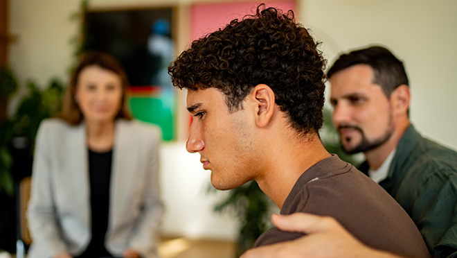 A teen sitting in a mental health professional&rsquo;s office with his father&rsquo;s arm wrapped around his shoulder in comfort.