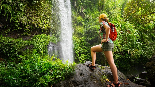 A woman in shorts and carrying a red backpack hikes near a waterfall in a densely forested area