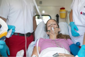  A young woman with long brown hair lies on a gurney in an ambulance. Her eyes are closed and she is wearing an oxygen mask and a blood pressure finger monitor. To either side of her, attending her, are a man and a woman whose faces are out of the picture frame. The woman is wearing a white uniform with a hospital cross and a pair of blue single-use gloves. The man is wearing a white polo, red pants, and blue single-use gloves. 
