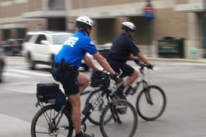 Two police officers on mountain bikes on a city street riding away and to the right of the camera. The background is dominated by a brick building with several large windows and entrances. They are crossing traffic and you can see a white SUV heading in their direction between them and the building.