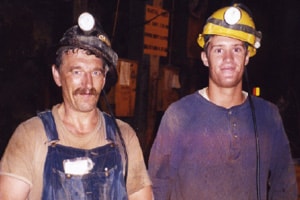 Two men look directly at the camera and grin. They are both very dirty. One man in overalls and t-shirt wearing a black miner’s hardhat with a light attached to the front looks to be about 50. Standing very close to this man is a younger man, probably in his twenties. He is wearing a dirty blue shirt and a yellow miner’s hard hat with a light attached to the front and a smaller one just above his right ear.