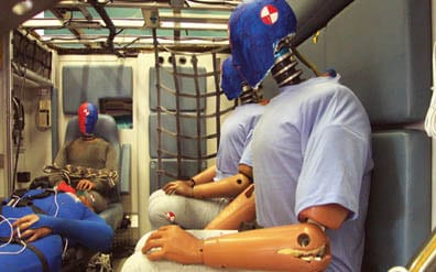 Photo of the inside of an ambulance simulator. 3 crash-test dummies have been positioned sitting upright in the ambulance crew seats. Another dummy lies on a gurney in front of them. The dummy in the foreground has a rip on its elbow from wear and tear. All the dummies and objects are in place and it is clear the crash simulation has not yet happened.