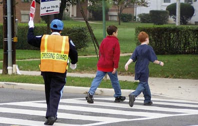 In this photo a woman in a blue uniform stands in the middle of a street crosswalk. She wears an orange and yellow pinafore with the words crossing guard written on it in large black letters. She is holding a stop sign in her left hand. 2 adolescent boys cross the street in front of her.