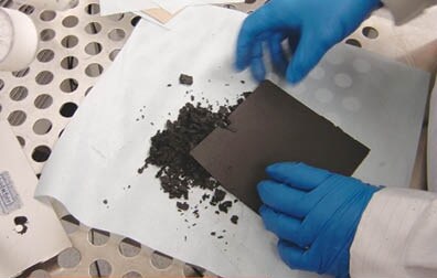 Close-up photo showing blue-gloved hands holding a crumbling black sheet of some sort of material. Under the hands is a pile of the material on a white cloth. Under the cloth is a metal sheet with large holes. In addition to the gloved hands there is also a bit of white lab coat showing.