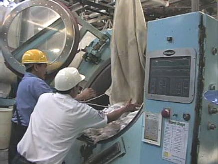 Exhibit #8. View showing two workers emptying laundry sling into tilted industrial washing machine.