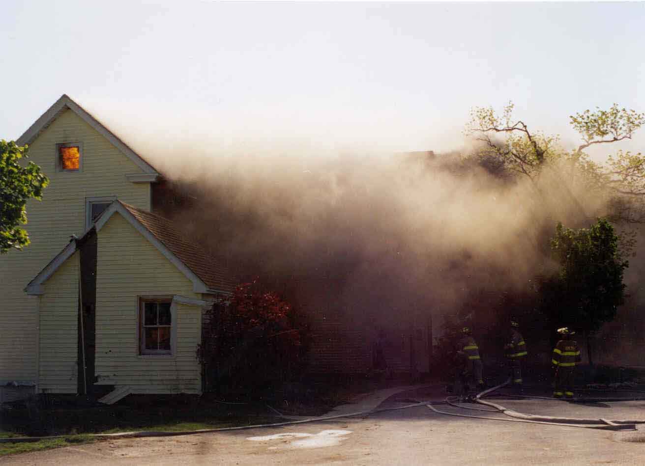 Photograph of the burning structure, with three fire fighters manning hoselines.