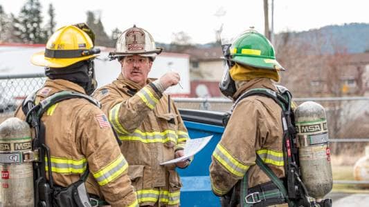 A Fire Chief holding a pen and paper in discussion with 2 firefighters on the scene of an emergency. Firefighting Concept