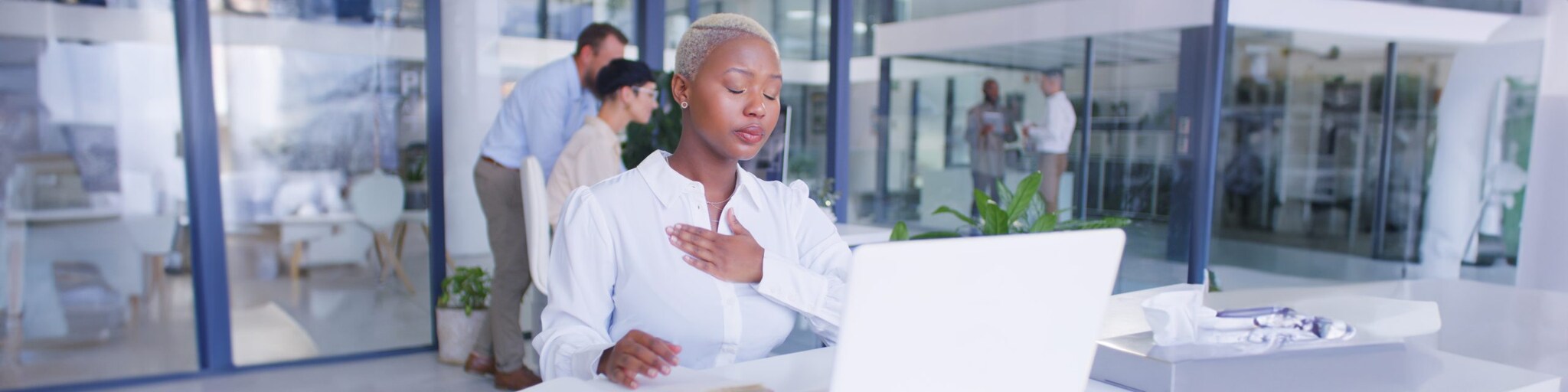African American woman with hand over heart, eyes closed, sitting at desk behind white laptop.