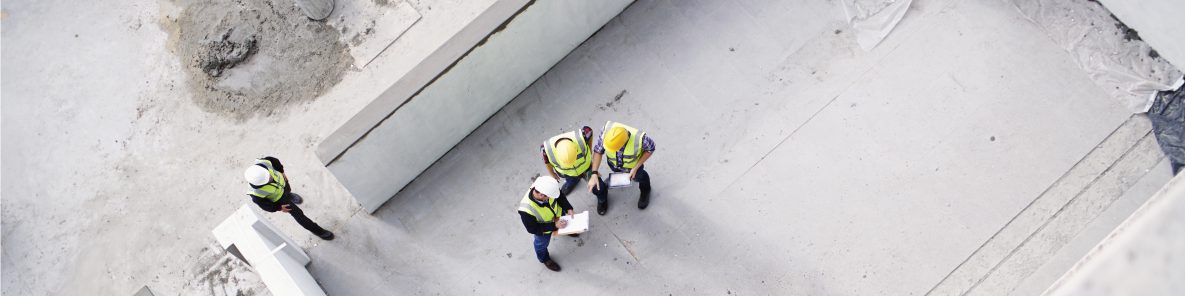 Three workers wearing hard hats and neon vests stand huddled over clipboards on cement platform, while a fourth worker enters the space.