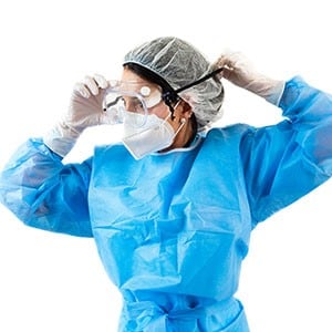 Healthcare worker wearing protective hospital gown, surgical gloves, hair net, respirator and putting protective eye wear on.