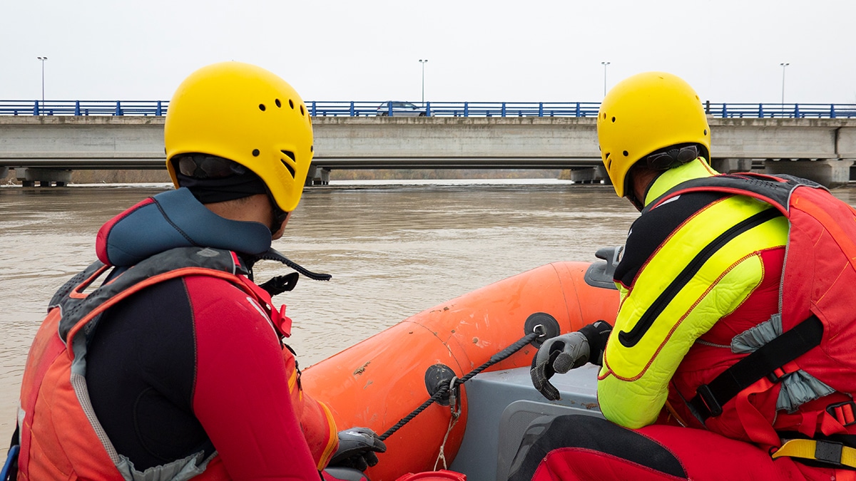two workers wearing a yellow helmet driving a boat on floodwater