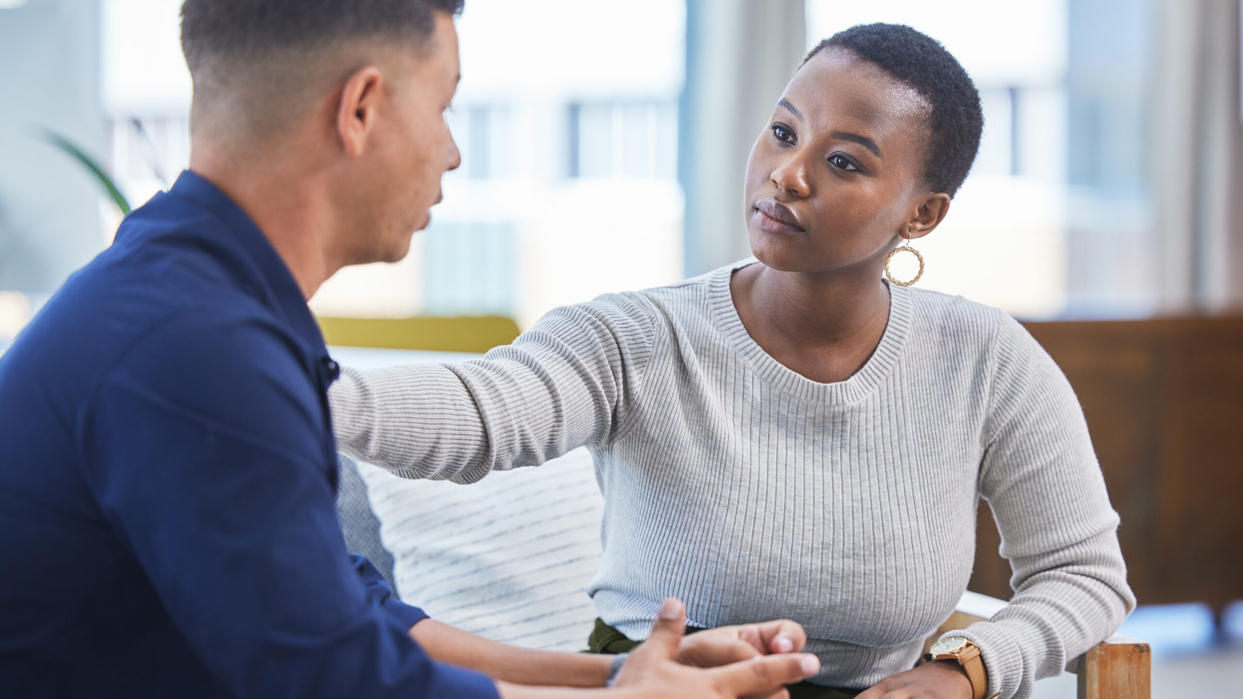 black woman in a gray sweater listening to and offering support to a man with his hands together, talking.