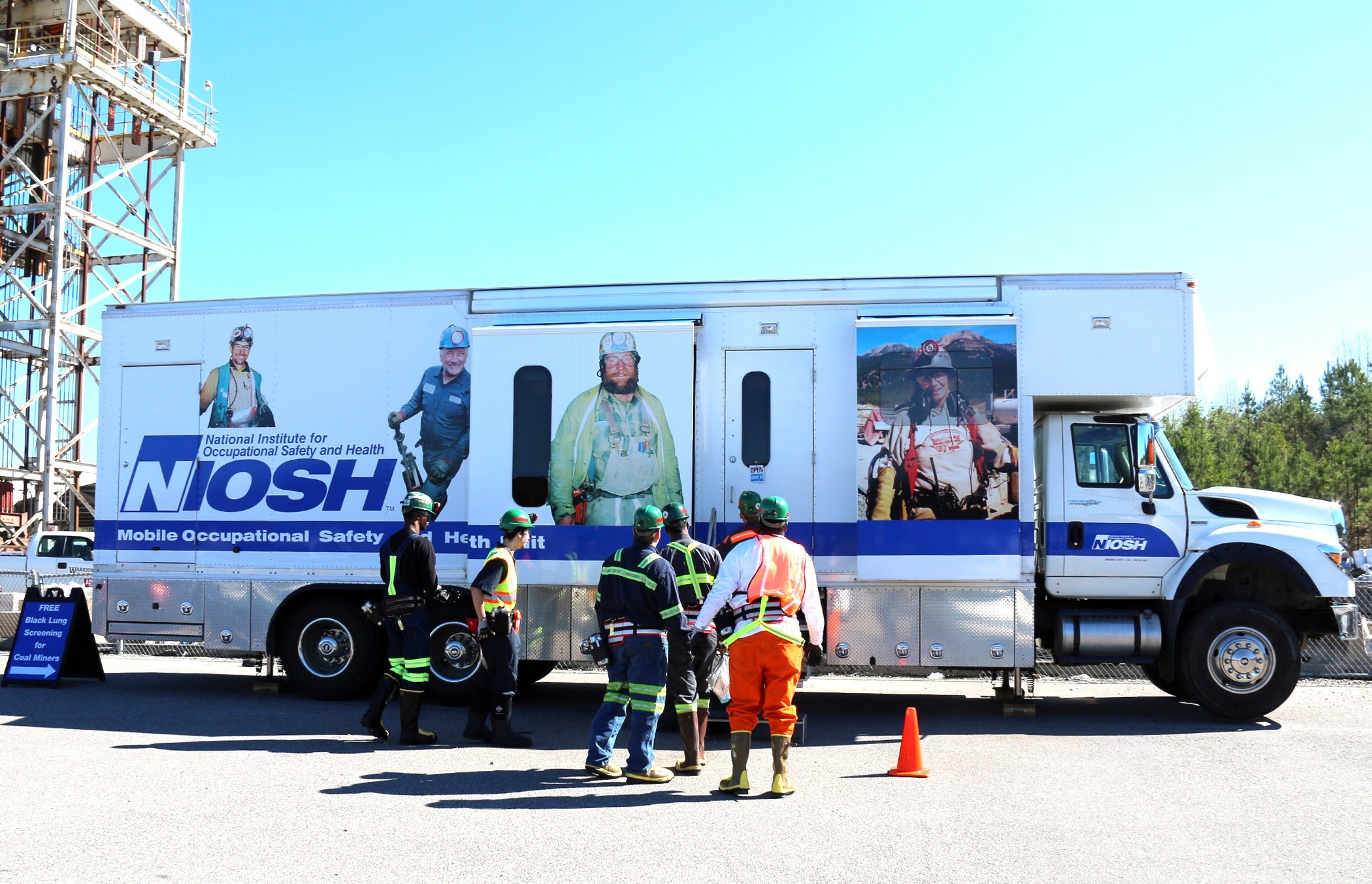 NIOSH’s Mobile Occupational Safety and Health Unit parked outdoors, with several workers in safety gear standing in front of the vehicle. The truck displays large images of miners and the NIOSH logo. A sign nearby advertises free black lung screenings for coal miners.