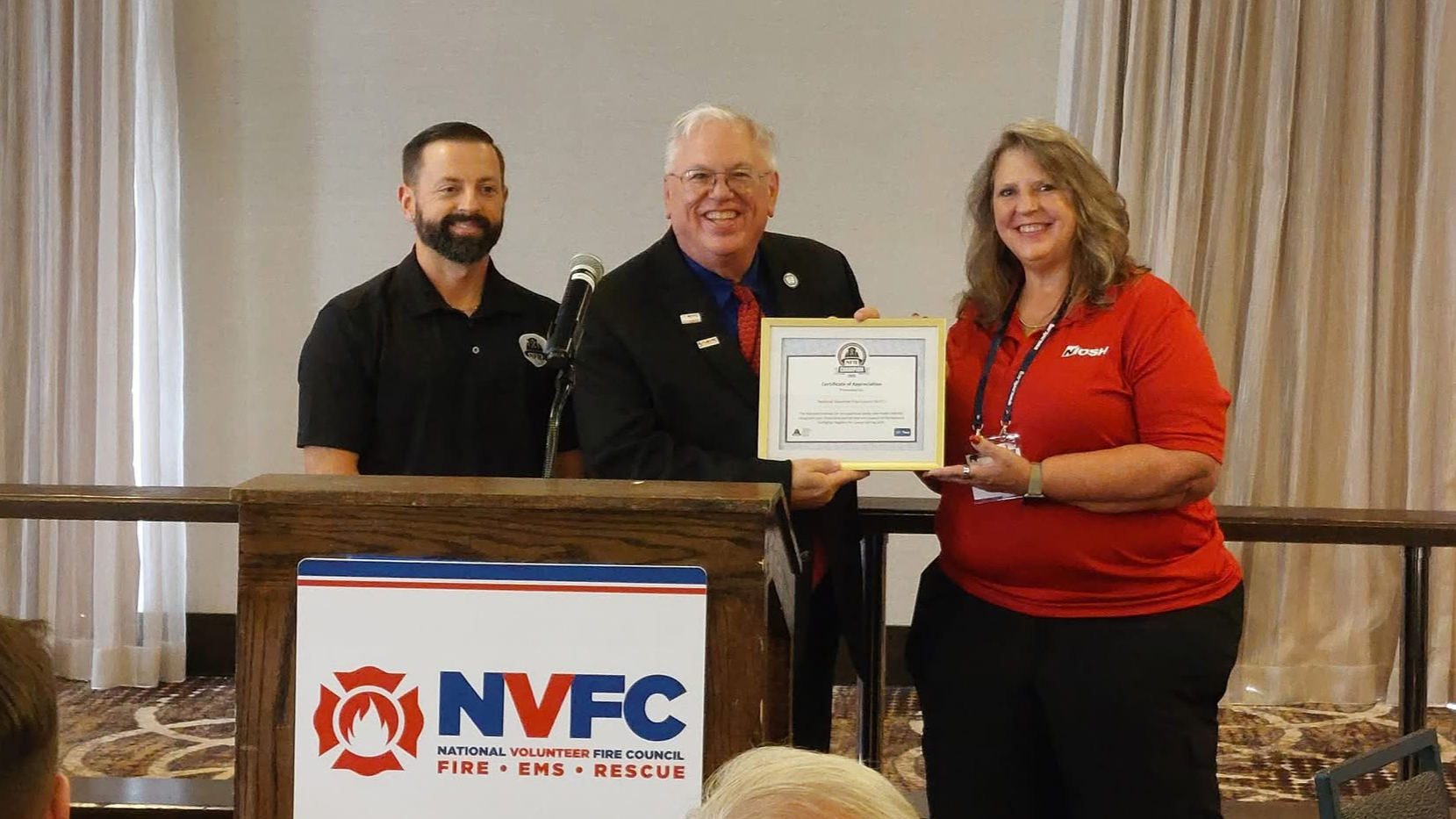 Two men and one woman stand behind podium with NVFC holding a certificate